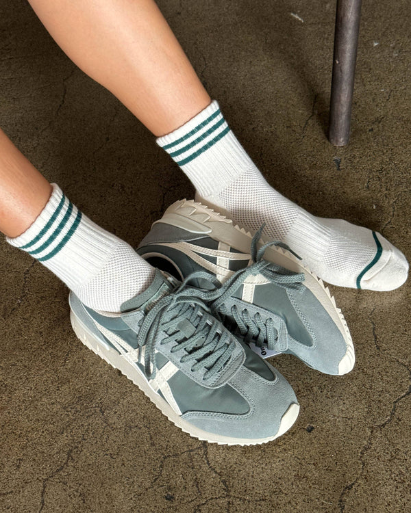 Person wearing gray sneakers and white socks with green stripes on a concrete floor.