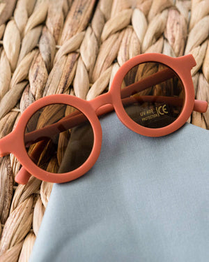 Round coral toddler-sized sunglasses with a reflective surface on a woven mat.