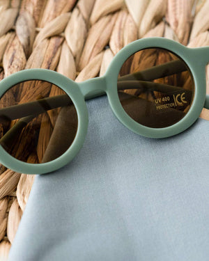 Round teal toddler-sized sunglasses with reflective lenses on a woven surface.