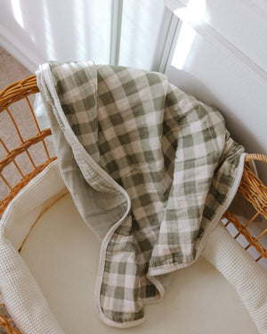 Green and beige gingham blanket draped over a wicker bassinet in front of a white wall