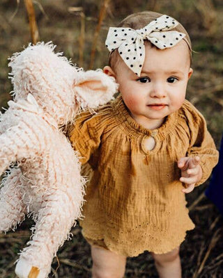 Baby in a mustard yellow outfit with a polka dot headband holding a plush pig toy outdoors.
