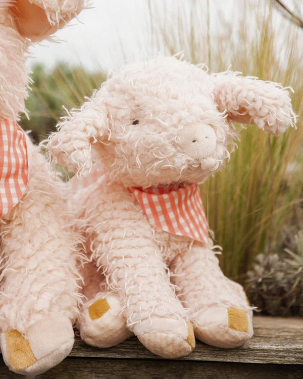 Fluffy pink pig with a red checkered scarf sitting on a wooden surface with grass in the background.
