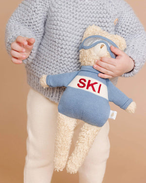 Child holding a teddy bear with 'SKI' on its shirt against a beige background