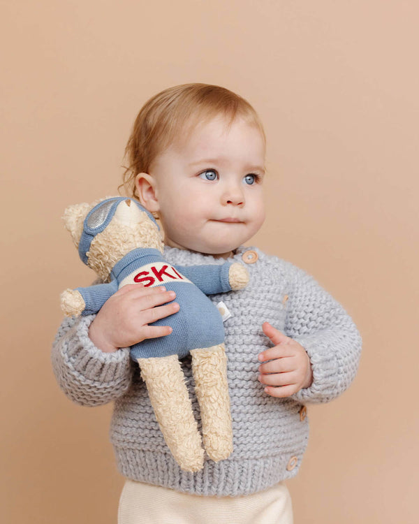 Child wearing a light blue knit sweater holding a teddy bear with goggles and 'SKI' text on a beige background