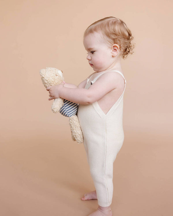 Child in a white romper holding a teddy bear against a beige background
