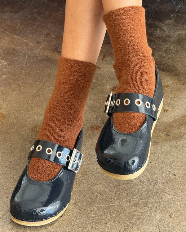Black clogs with strap details worn with brown socks on a concrete floor.