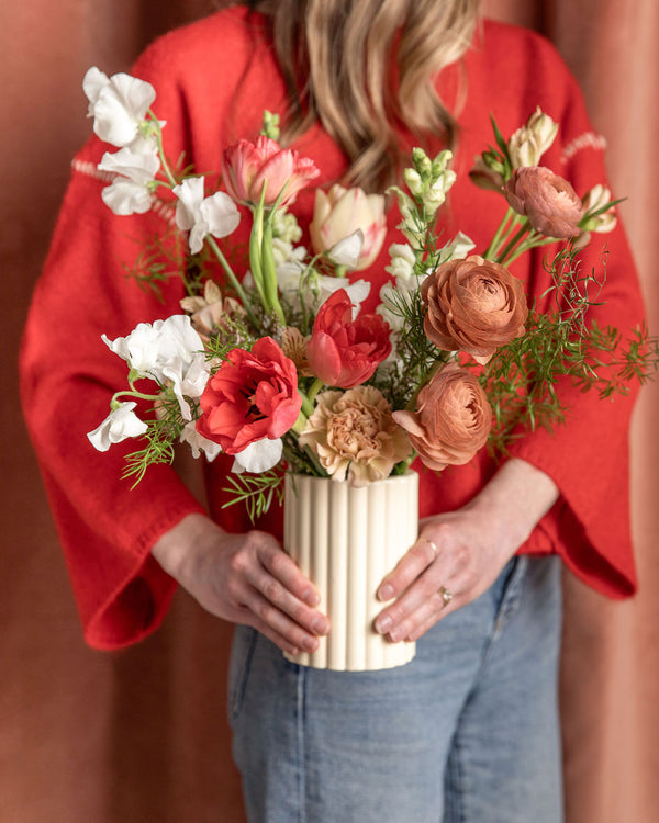 Love Story Valentine’s Day arrangement held by a person in jeans and a red sweater, featuring tulips, ranunculus, snapdragons, and sweet peas in a cream ridged vase.