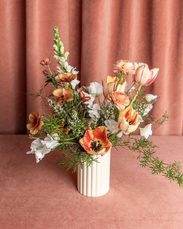 Love Story Valentine’s Day vase arrangement styled on a rosy backdrop, designed with tulips, ranunculus, snapdragons, sweet peas, and airy greenery in a cream ceramic vase.