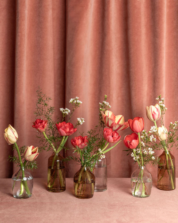 Assortment of Lovebug Valentine’s Day bud vases featuring tulips, waxflower, and soft greenery in clear and amber glass.