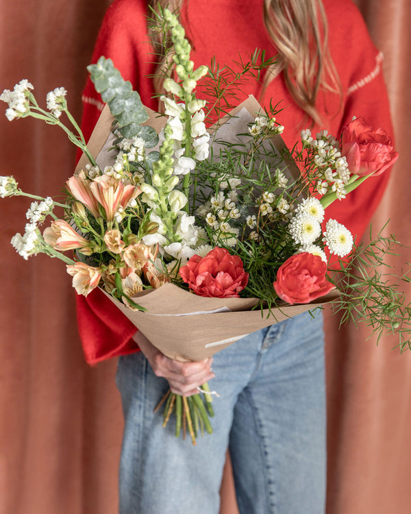 Natural Valentine’s Day bouquet with peach and coral blooms held by a person in jeans and a red sweater, designed by Landfall Floral Co.