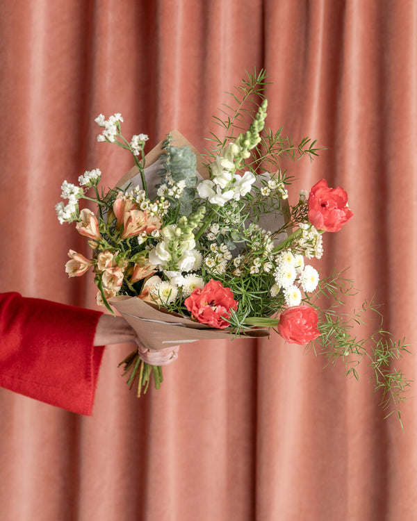 Valentine’s Day Signature Bouquet “The Luckiest,” featuring coral tulips, white snapdragons, and seasonal greenery against a soft pink backdrop.